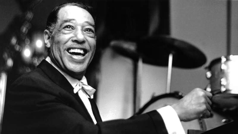 A black and white photo of jazz musician Duke Ellington smiling at the camera while sitting at a piano