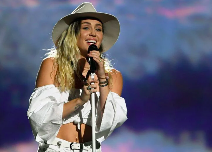 Singer Miley Cyrus smiles while holding a microphone and wearing a cowboy hat